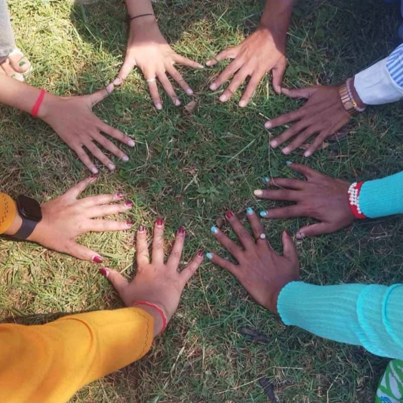 Eight hands with colorful nail polish are arranged in a circle on a grassy ground, with shadows cast by sunlight.