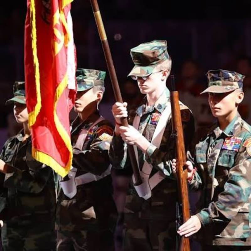 Four young people in camouflage uniforms stand at attention, holding a flag and a rifle, with a blurred crowd in the background.
