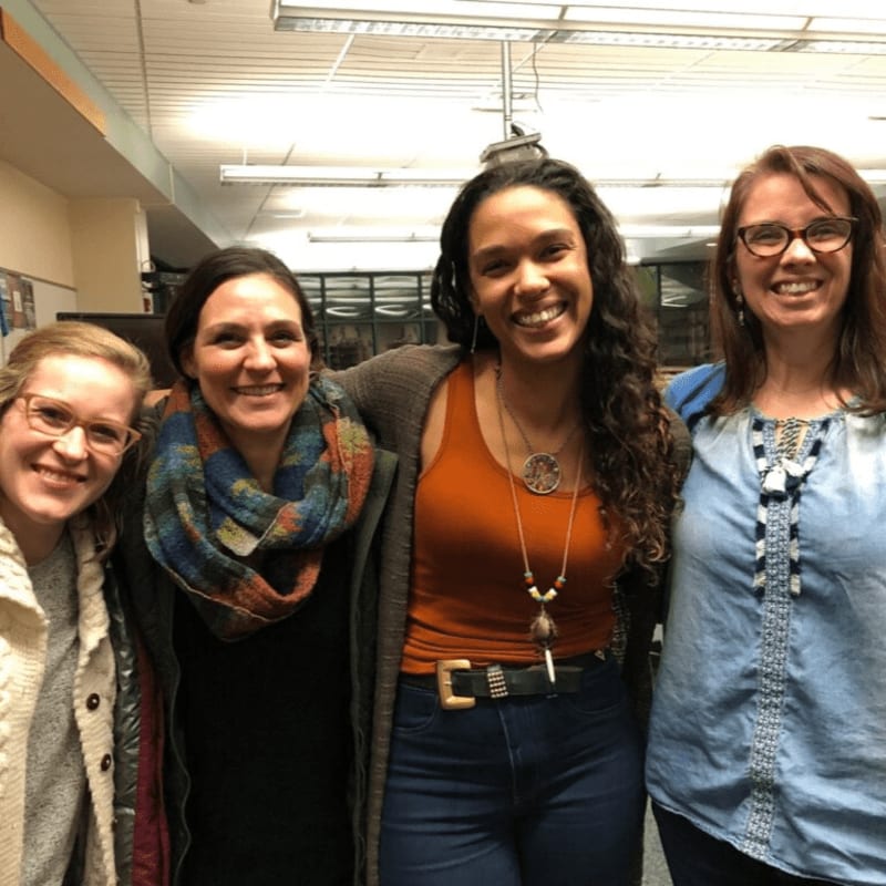Four smiling women pose together in the foreground, with a blurred indoor background of fluorescent lighting and what appears to be office or classroom walls.