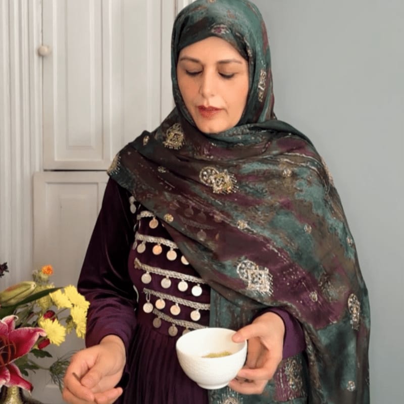 A woman in a purple dress and patterned hijab holds a cup of tea in the foreground, with a bouquet of flowers to her left and a white cabinet in the background.