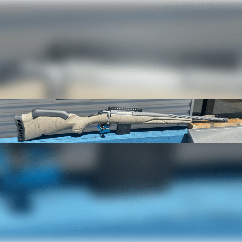 A tan speckled rifle rests on a blue surface in the foreground, with a blurred building in the background.
