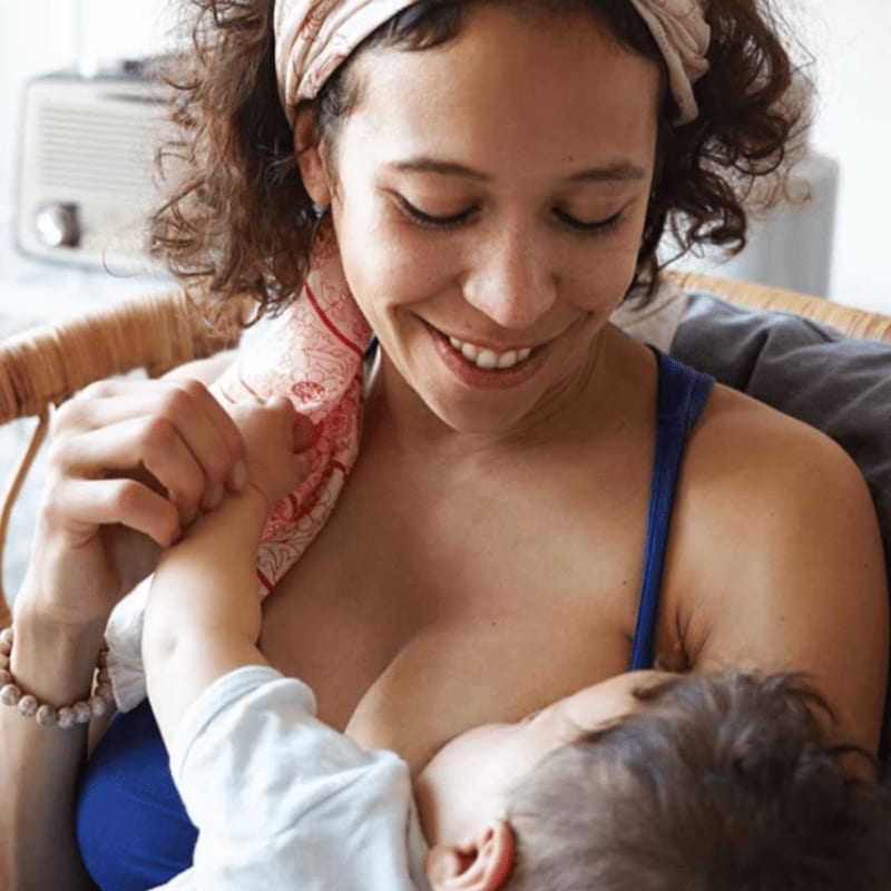 A smiling mother with a bandana in her curly hair breastfeeds her baby, with a wicker chair and a radio visible in the soft-focus background.