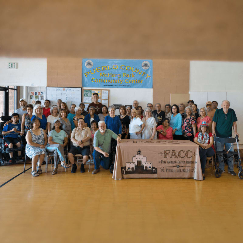 A large group of people, some in wheelchairs, pose for a photo in front of a banner and a table with a logo in a gymnasium.