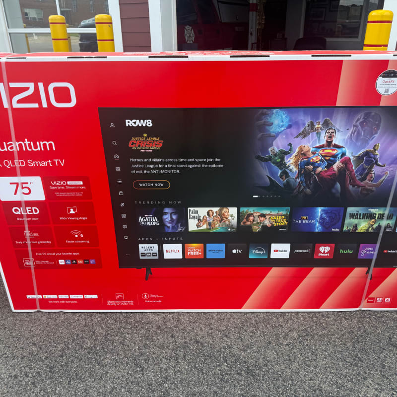 A large VIZIO QLED Smart TV box with a superhero movie displayed on its screen sits on a gray surface, with yellow bollards and a building visible in the background.
