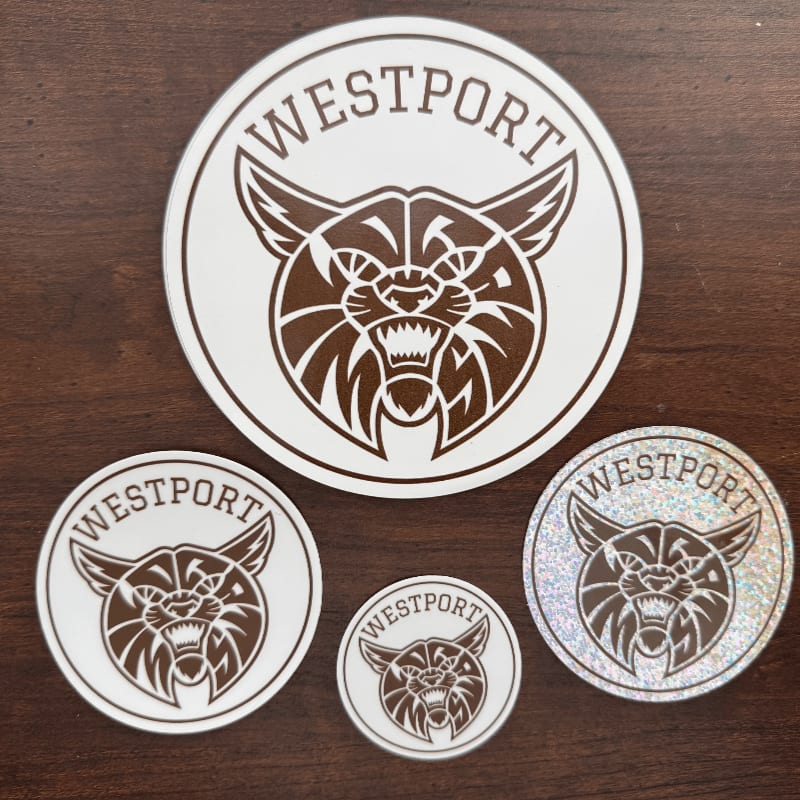 Four Westport bobcat stickers of varying sizes are arranged on a dark wooden surface.