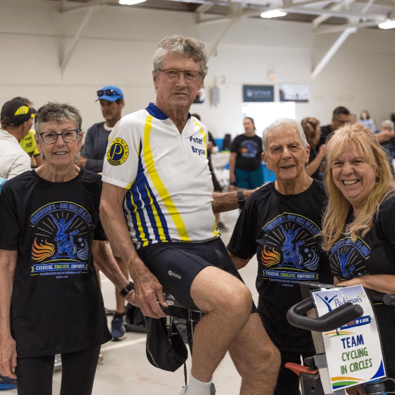 Four smiling people, two men and two women, pose in the foreground with stationary bikes, while a blurred background shows a gymnasium with other participants.