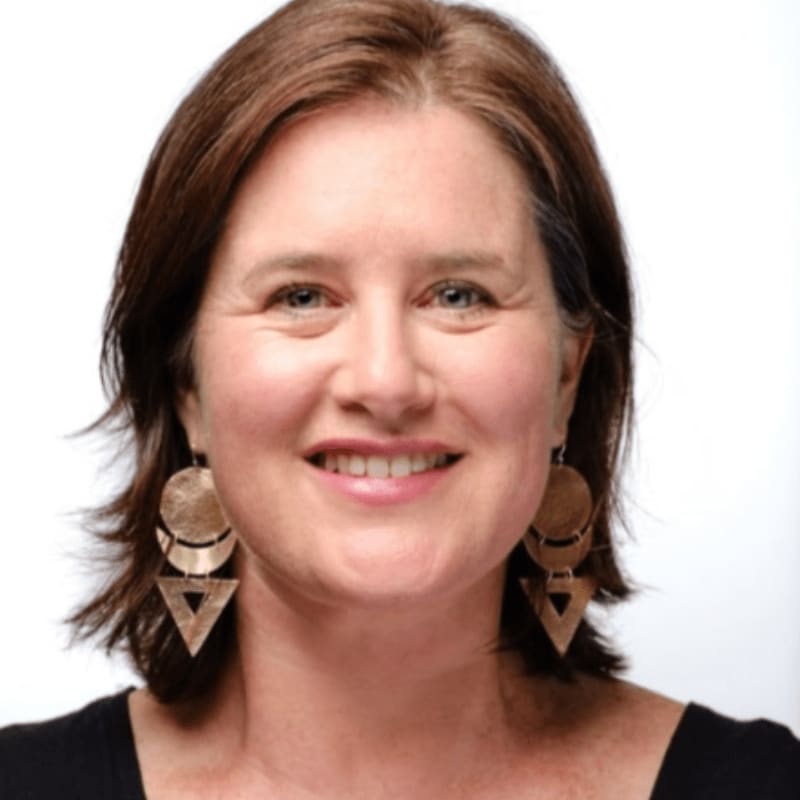 A woman with brown hair and large, geometric earrings smiles at the camera against a plain white background.