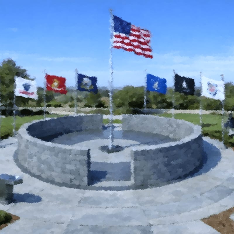 A circular stone monument with several flags flying above it stands in the foreground, while a clear blue sky and distant green hills form the background.