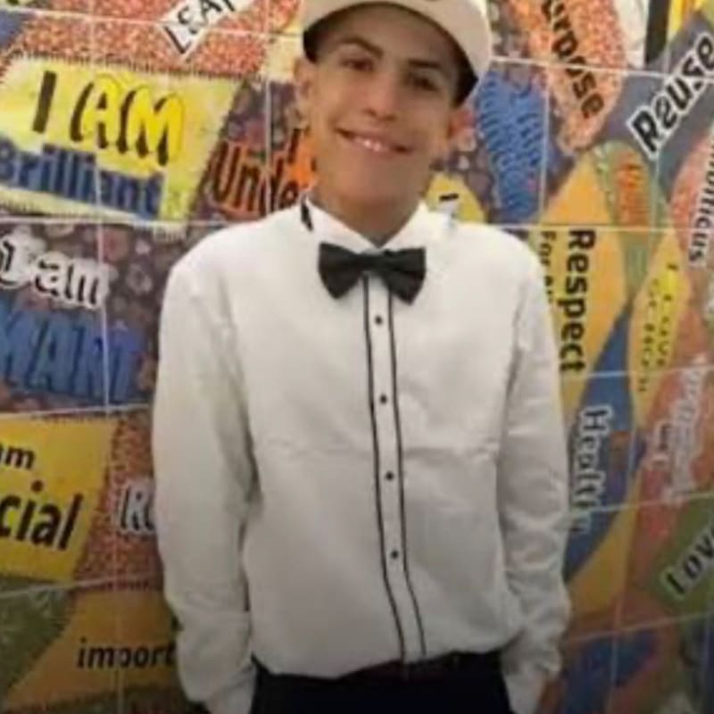 A young man in a white shirt and bow tie smiles in front of a colorful, text-covered wall.