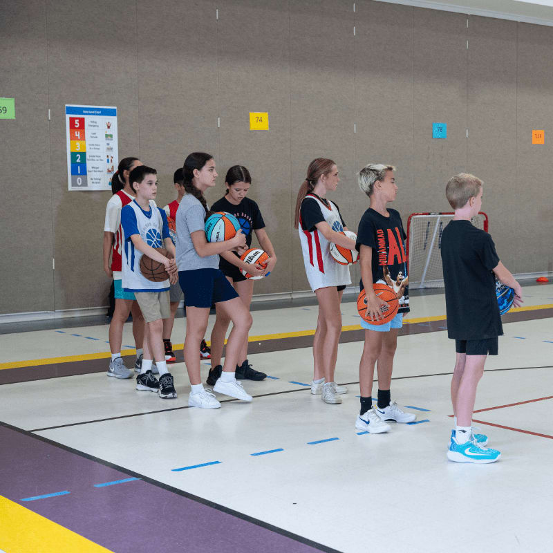 Children in athletic wear stand in a line holding basketballs on a polished gym floor with a colorful wall and a hockey net in the background.