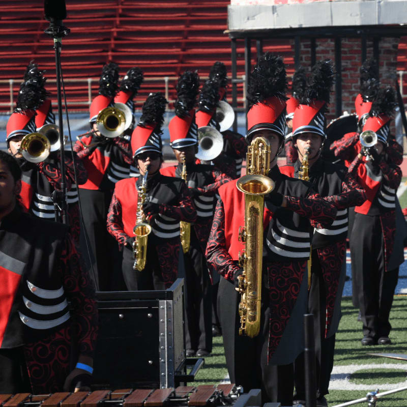 A marching band in red and black uniforms plays instruments on a field in front of stadium bleachers.