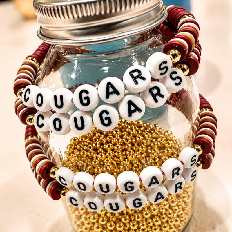 Two beaded bracelets spelling "Cougars" in white letters with gold accents are wrapped around a glass jar filled with gold beads, with a blurred background.