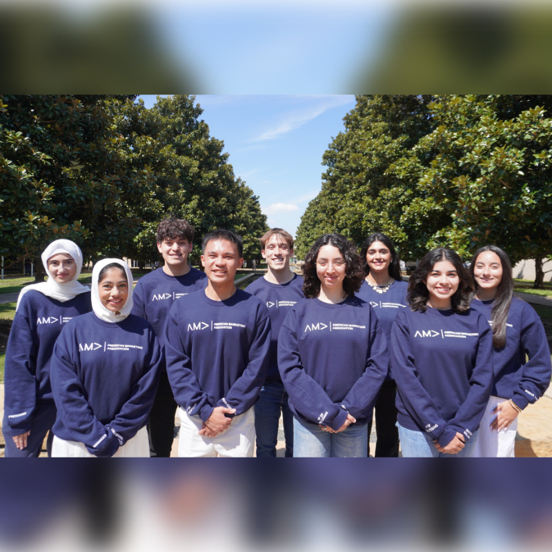 A group of nine young people in matching navy sweatshirts stand in a park with manicured trees lining a path behind them.