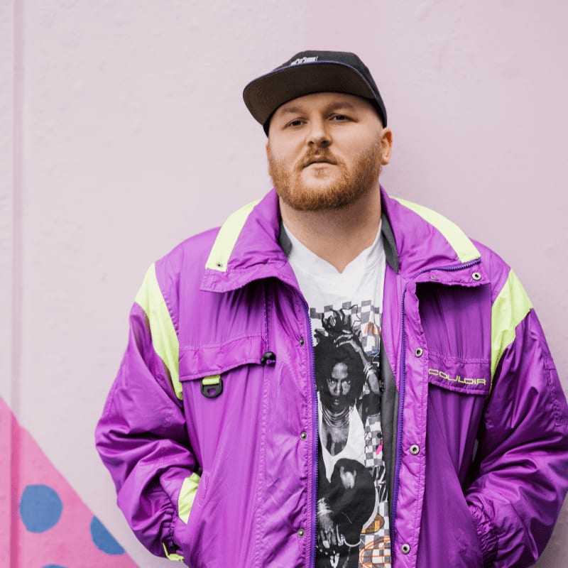A man in a purple jacket and a Bob Marley t-shirt stands in front of a pink wall with blue polka dots.