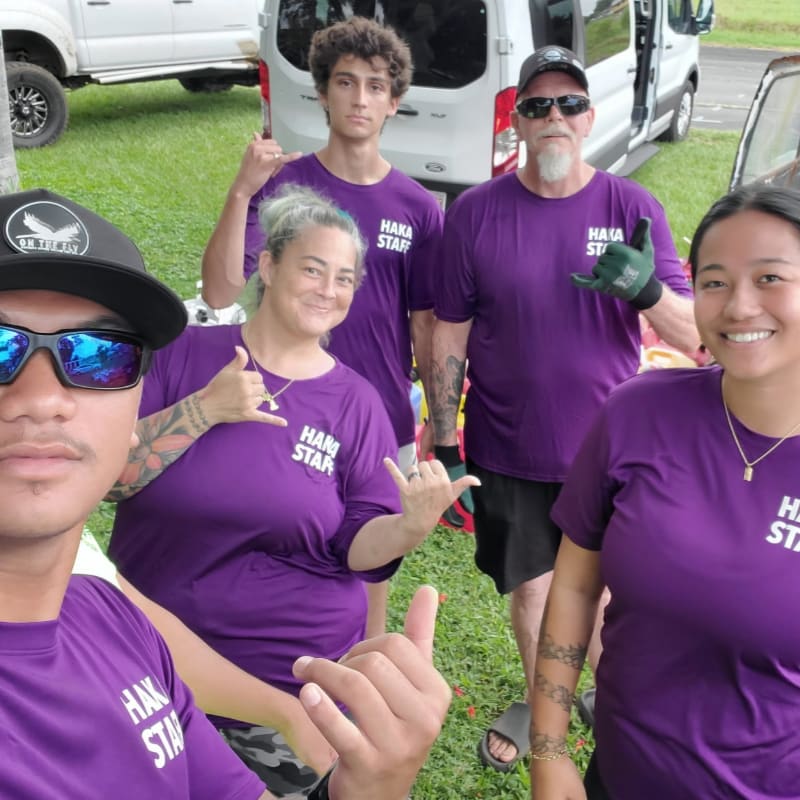 Five people in purple shirts with "HAKA STAFF" printed on them pose for a selfie in front of white vans and a grassy area.