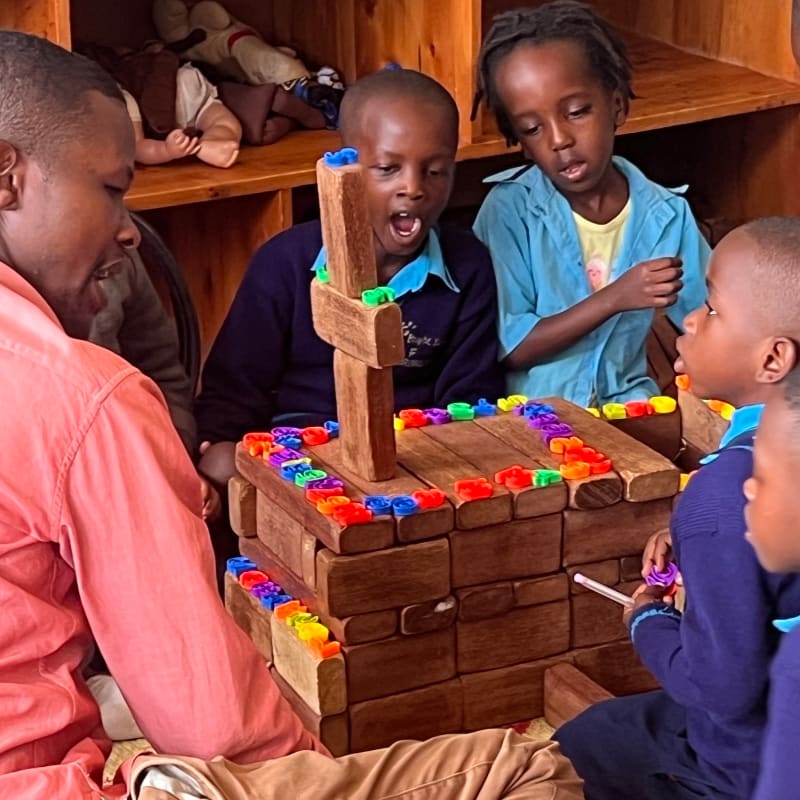 A man in a pink shirt and children in school uniforms engage with a wooden block structure adorned with colorful pieces in the foreground, while shelves filled with dolls and toys are visible in the background.