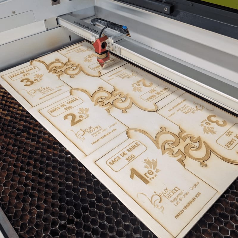 A laser cutter engraves intricate designs and text onto a wooden board in the foreground, while the honeycomb-patterned bed of the machine forms the background.