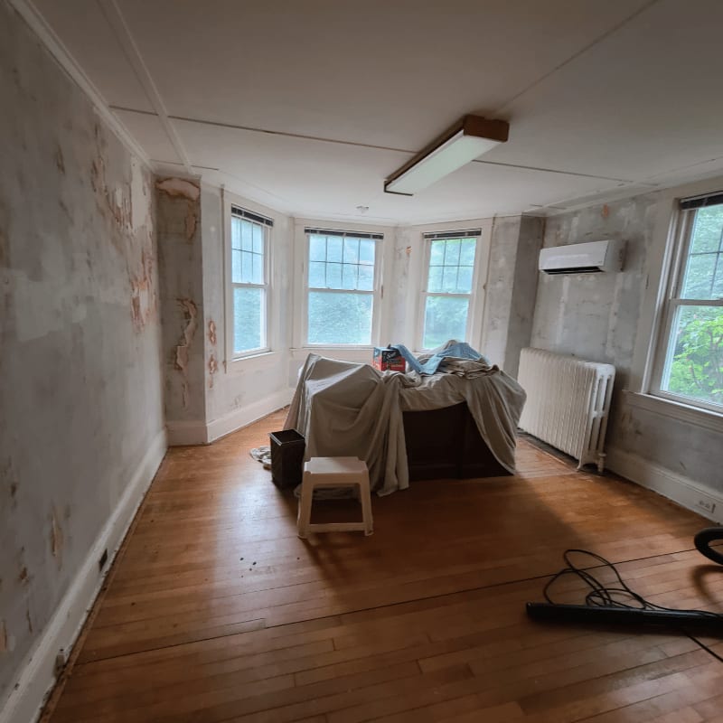 A small stool and trash can sit on the wooden floor in the foreground, while a large bed covered in a sheet and bay windows with blinds dominate the background.