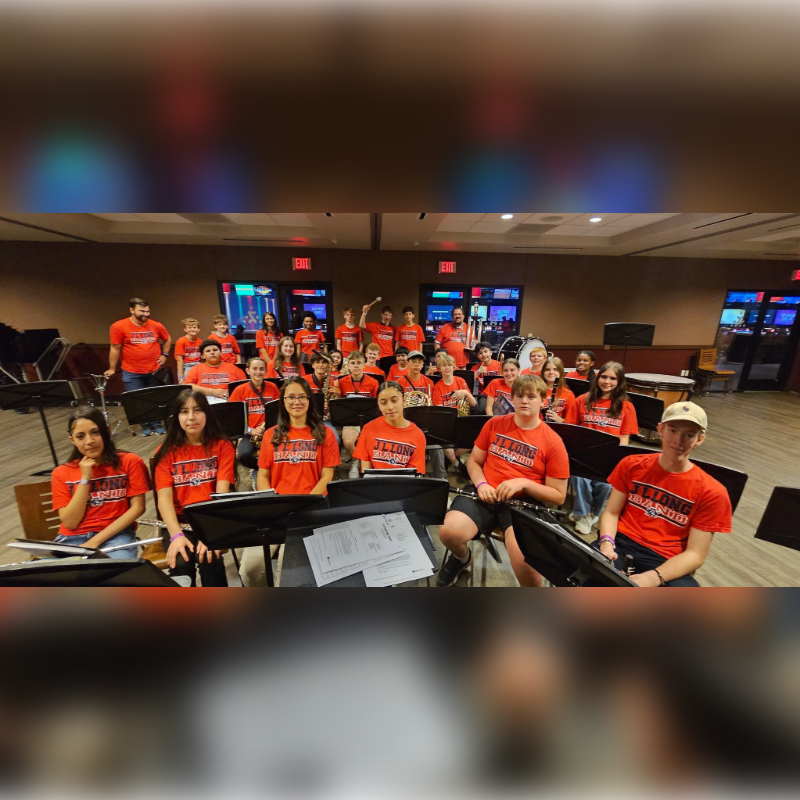 A group of young musicians in red shirts are seated with their instruments in the foreground, while a blurred background shows a hotel lobby with glowing screens.