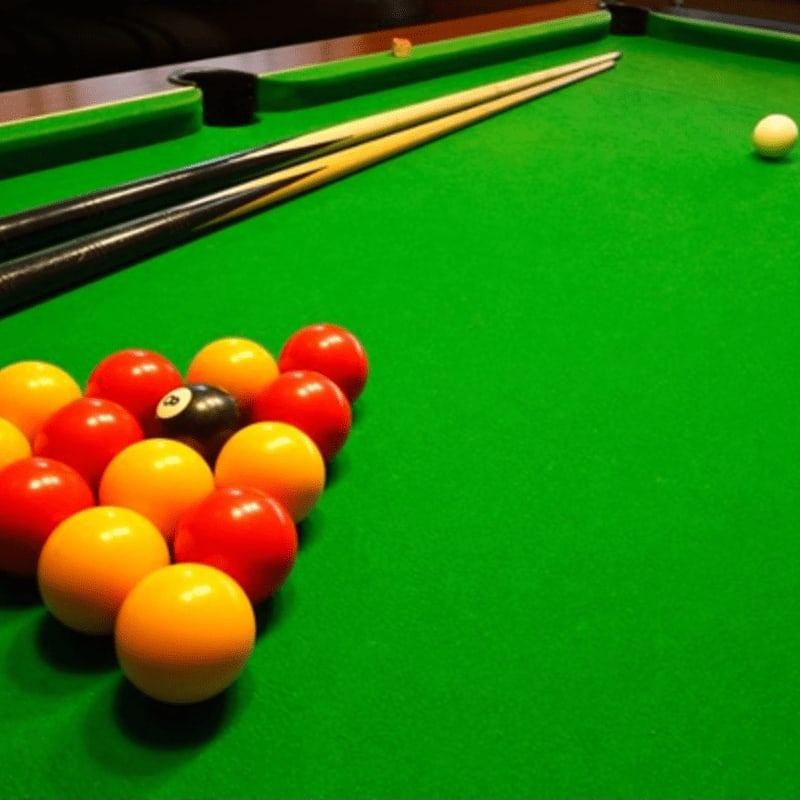 A rack of colorful billiard balls sits on a green felt table in the foreground, with two cue sticks resting diagonally behind them and a single white ball in the background.
