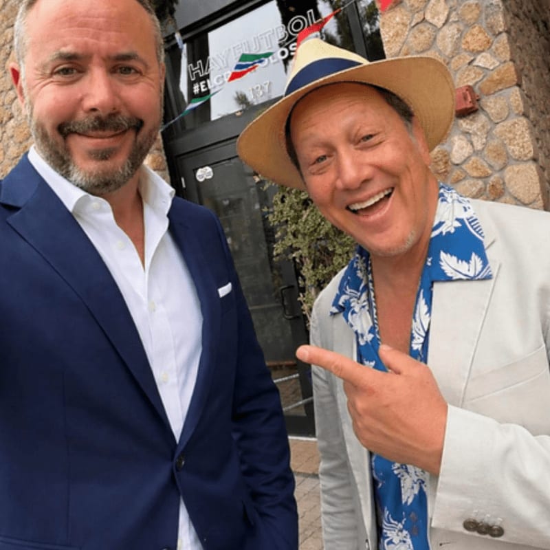 Two smiling men, one in a suit and the other in a straw hat and patterned shirt, pose in front of a stone building with a sign that reads "HAY FUTBOL".