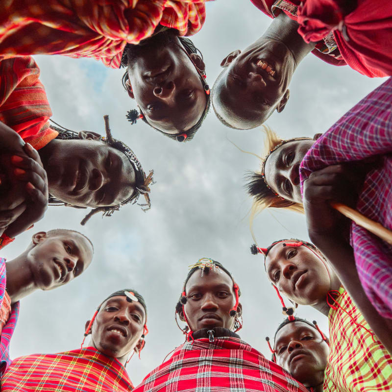 A group of Maasai men in traditional red and yellow checkered clothing form a circle, looking down from a low angle against a cloudy sky.