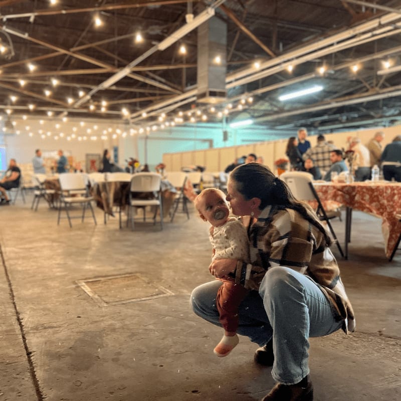 A woman in jeans and a plaid jacket holds a baby in her arms in the foreground, while the background shows a large, dimly lit hall with tables, chairs, and strings of lights.