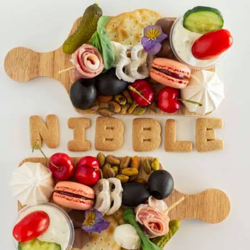 Two wooden boards filled with a colorful assortment of snacks, including macarons, olives, and pickles, are arranged around the word "NIBBLE" spelled out in cookie letters against a white background.