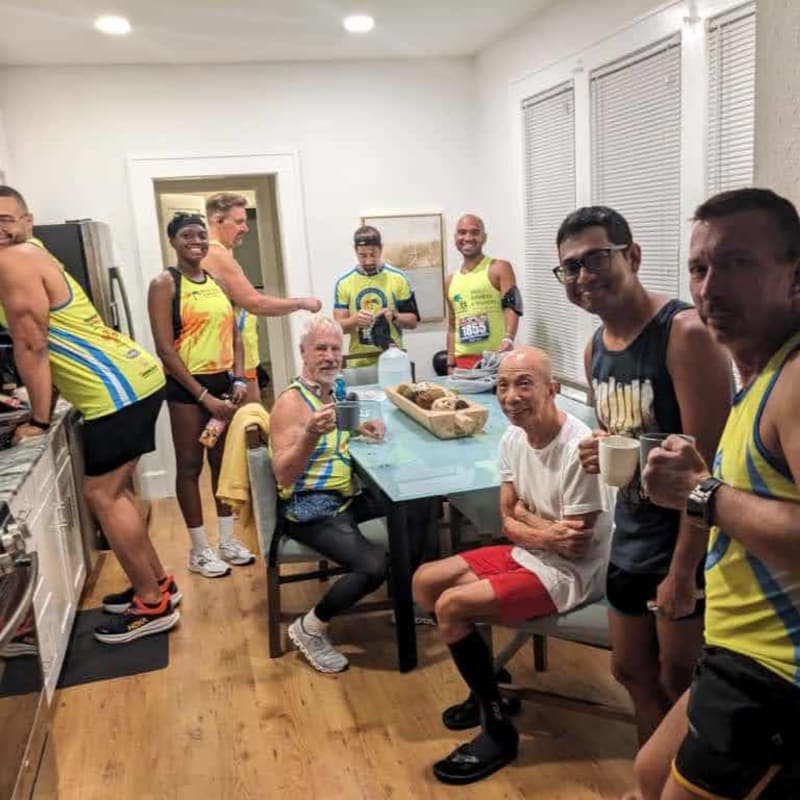 A group of runners in athletic wear gather around a table in a kitchen, with a doorway and cabinets visible in the background.