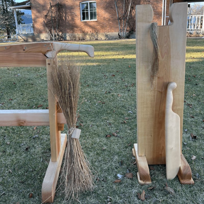A wooden broom leans against a rustic wooden stand in the foreground, with a brick house and bare trees in the background.