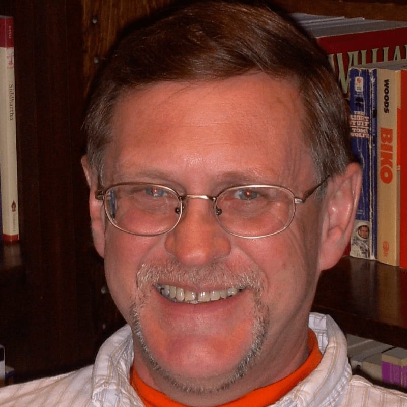 A smiling man with glasses and a goatee is in the foreground, with a bookshelf filled with books in the background.