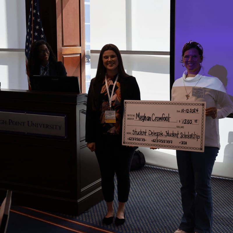 Two women stand holding a scholarship check in front of a podium with an American flag, while a third woman sits at a laptop in the background.