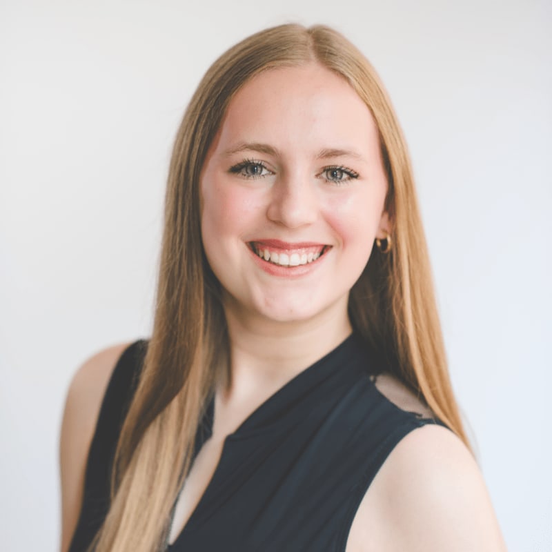A young woman with long blonde hair and a black top smiles at the camera against a plain white background.