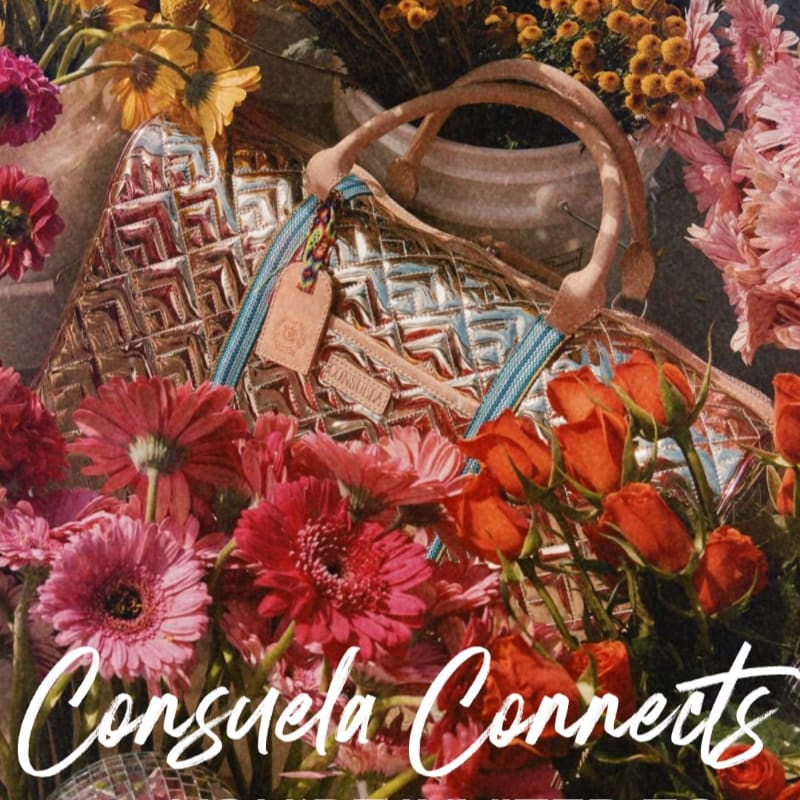A metallic patterned tote bag rests amongst a vibrant bouquet of pink and orange flowers, with a blurred background of more floral arrangements.