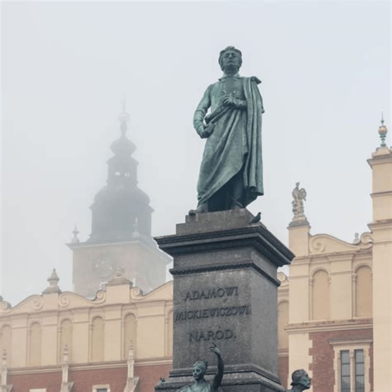 A bronze statue of Adam Mickiewicz stands on a pedestal in the foreground, with a hazy view of historic buildings in the background.