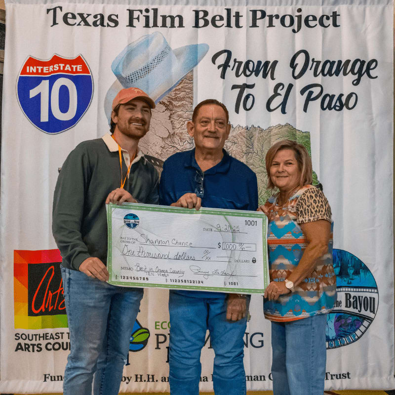 Three people stand in front of a banner for the Texas Film Belt Project, holding a large novelty check.