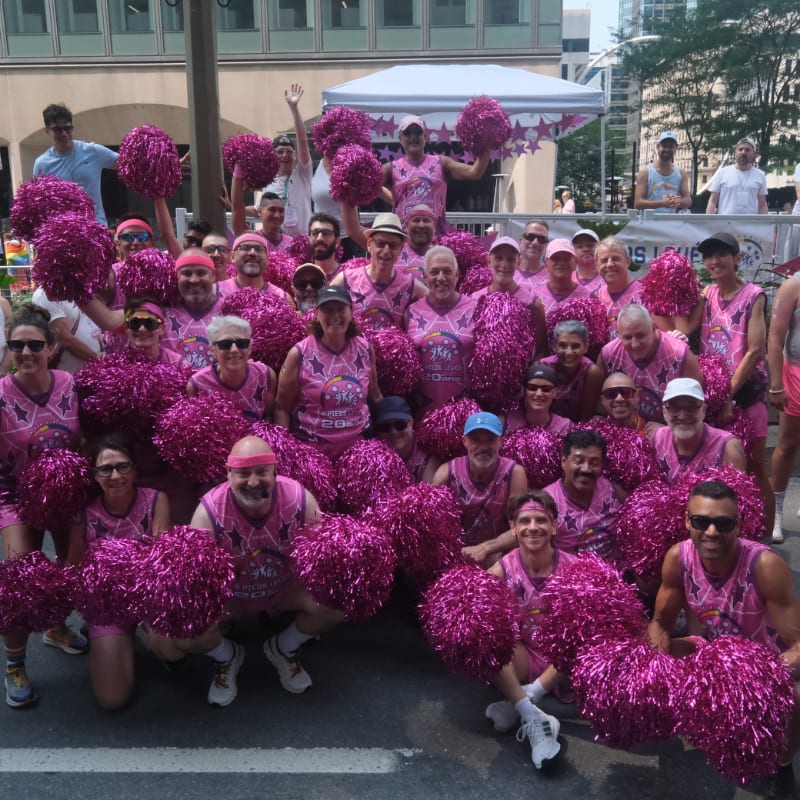 A group of people in pink shirts and holding pink pom-poms pose for a photo in front of a white tent and a blurred cityscape.