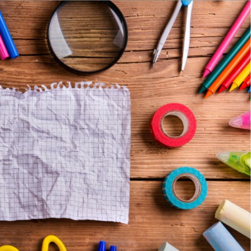 A crumpled piece of graph paper lies on a wooden surface alongside a magnifying glass, colored pencils, a compass, and rolls of tape.