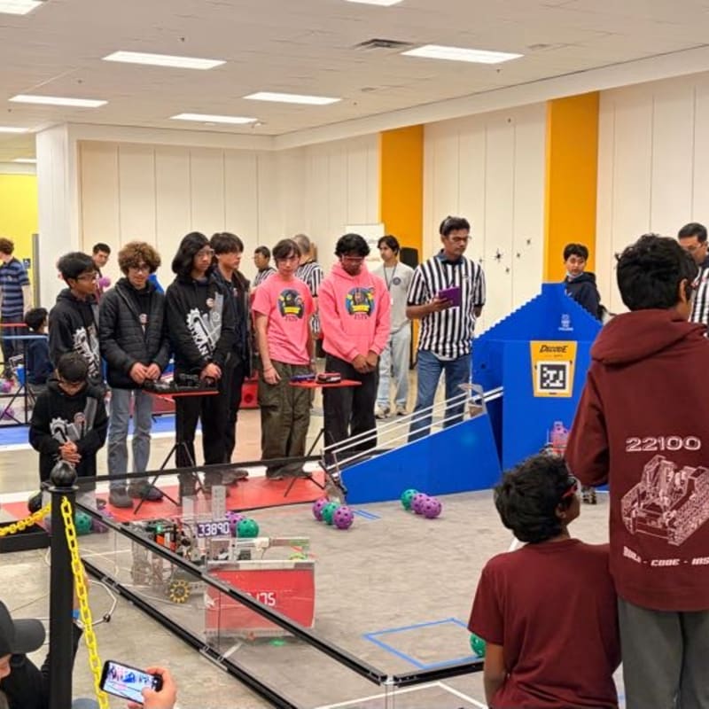In the foreground, students in casual attire observe a robotics competition with colorful balls on the floor, while in the background, a referee in a striped shirt stands near a blue structure.