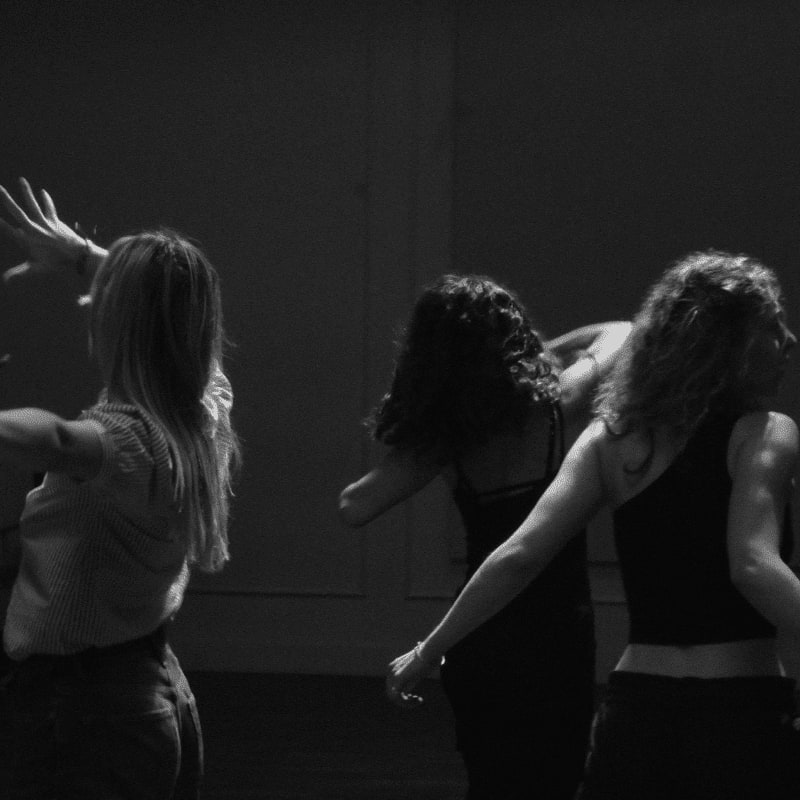 Three women with their backs to the viewer dance in a dimly lit room.