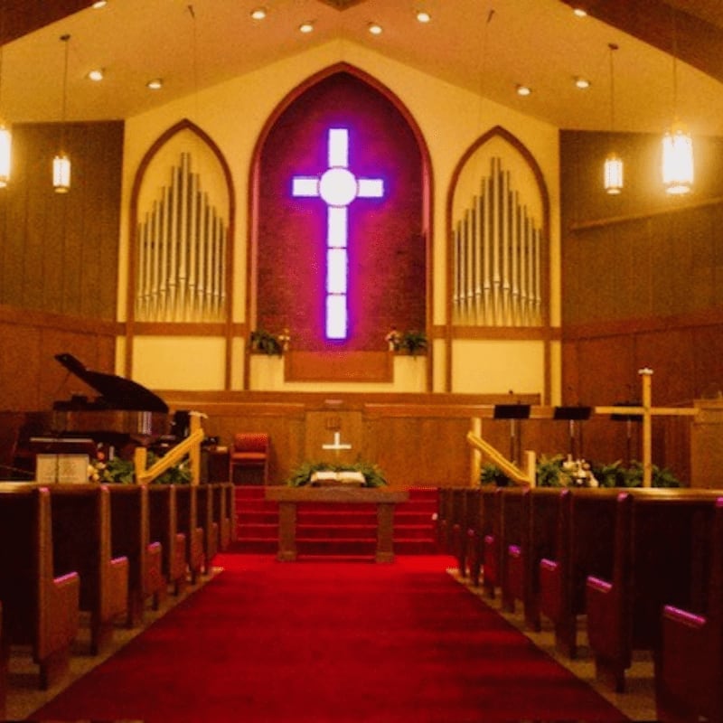 Rows of wooden pews line a red carpet leading to an altar with an open bible, while a large, illuminated cross and pipe organs adorn the background of the church.