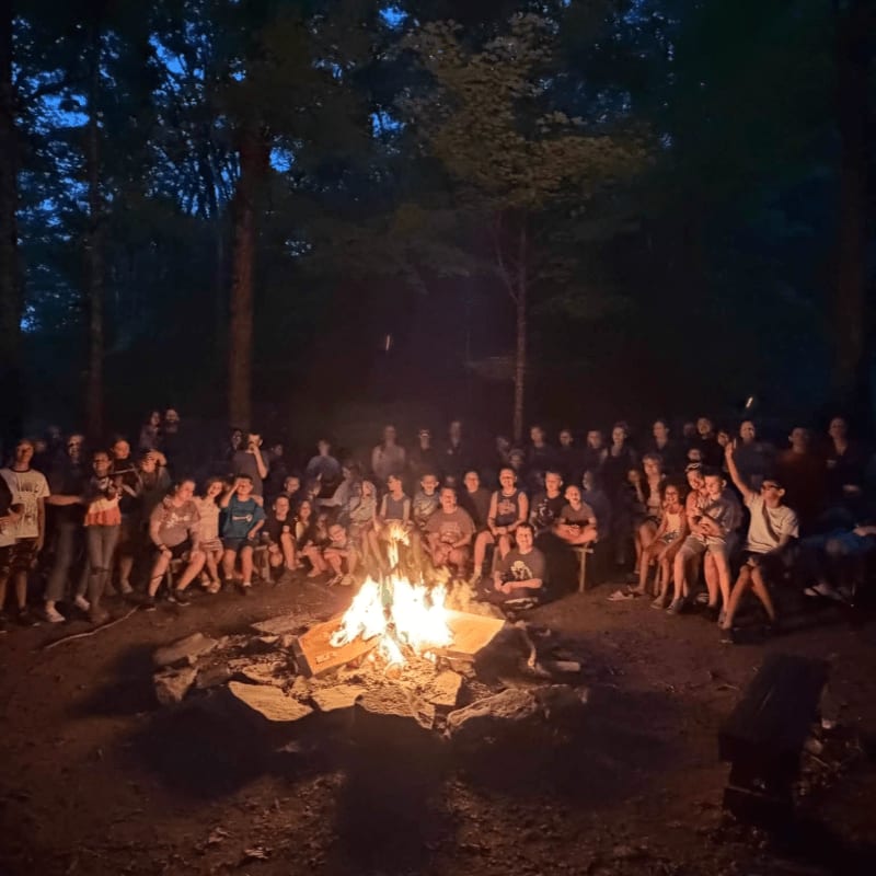 A group of people sit around a crackling campfire in a dark forest clearing.