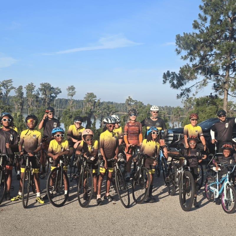 A group of children and adults in cycling gear stand with their bicycles in the foreground, while a body of water and trees form the background under a clear blue sky.