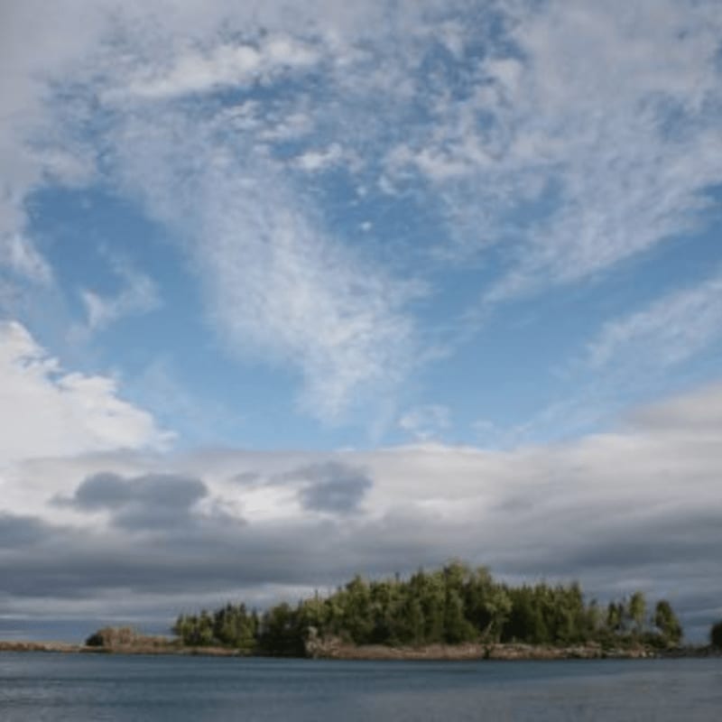A small, tree-covered island sits on calm blue water under a vast sky filled with wispy clouds.