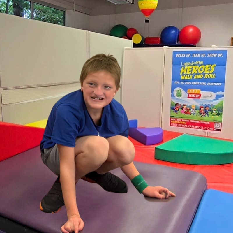 A young boy in a blue shirt and grey shorts crouches on a purple mat in the foreground, while a colorful poster and various exercise balls are visible in the background.