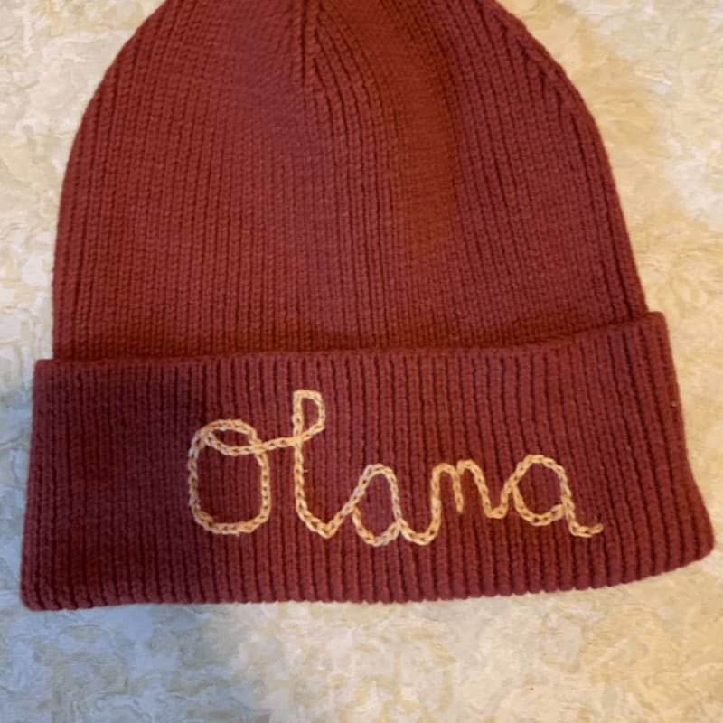 A maroon knit beanie with "Olana" embroidered in beige yarn rests on a textured white surface.