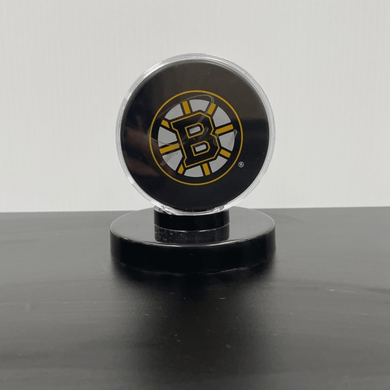 A Boston Bruins puck sits on a black stand in the foreground, with a plain white background behind it.