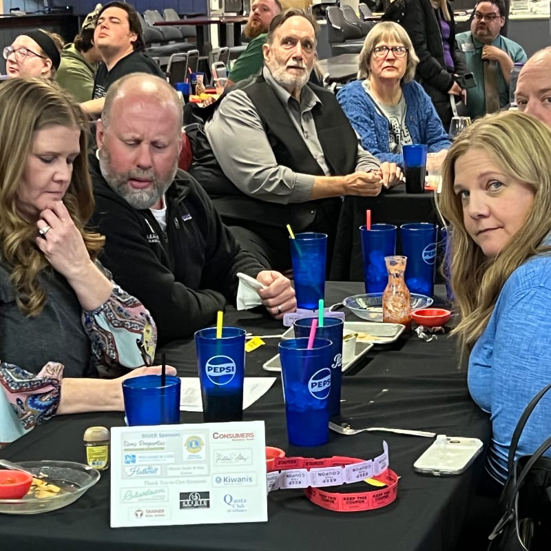 People are seated at a table with blue cups and raffle tickets in the foreground, while others are visible in the blurred background of a gathering.