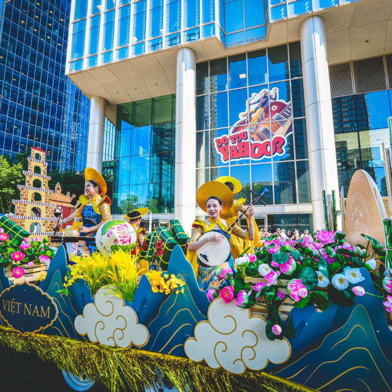 A vibrant parade float with performers in traditional attire and floral decorations moves past a modern skyscraper with a "Do You Yahoo?" sign.