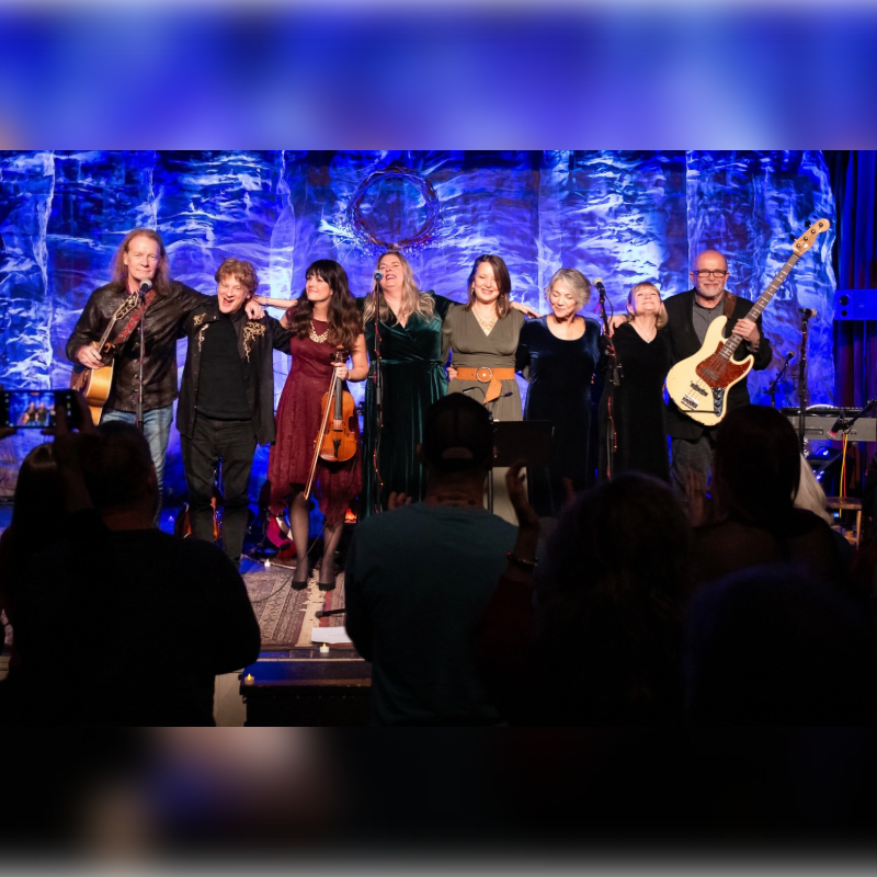 A band of seven musicians stands on a stage with a textured blue backdrop, while an audience watches from the foreground.
